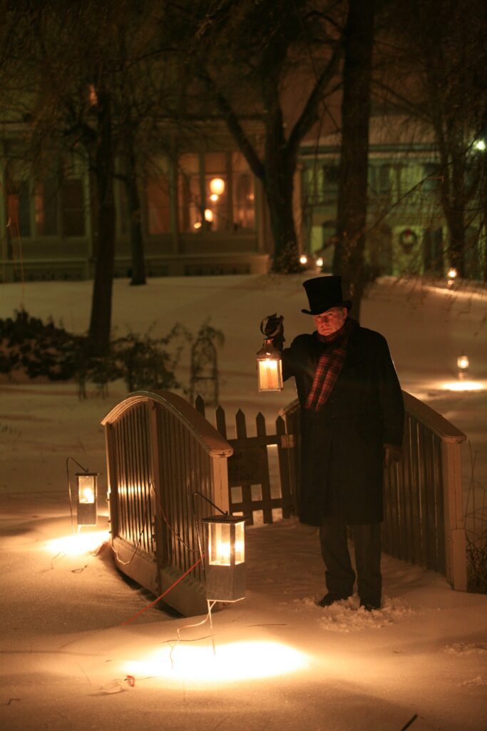 A man in winter garb, plus top hat holds a lantern a lot inviting guests up the path to Villa Louis as part of his employment. 